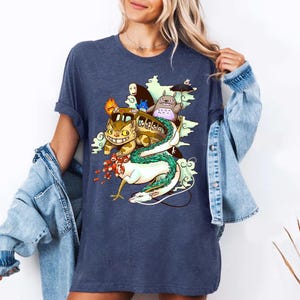 K&ouml;nnte beinhalten: Ein marineblaues T-Shirt mit einer farbenfrohen Illustration von Charakteren aus einem beliebten Animationsfilm. Das Design zeigt ein busf&ouml;rmiges Wesen, eine gro&szlig;e katzenartige Figur und einen langen wei&szlig;en Drachen. Das Shirt ist ein l&auml;ssiger Stil.