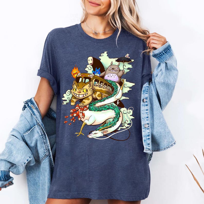 K&ouml;nnte beinhalten: Ein marineblaues T-Shirt mit einer farbenfrohen Illustration von Charakteren aus einem beliebten Animationsfilm. Das Design zeigt ein busf&ouml;rmiges Wesen, eine gro&szlig;e katzenartige Figur und einen langen wei&szlig;en Drachen. Das Shirt ist ein l&auml;ssiger Stil.