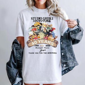 May include: White t-shirt featuring a colorful graphic of Studio Ghibli characters. The shirt commemorates 40 years with text that reads "STUDIO GHIBLI", "40 YEARS", and "THANK YOU FOR THE MEMORIES". The design includes Japanese text and the years 1985 and 2025.