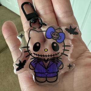 May include: A clear acrylic keychain featuring a cartoon cat character dressed in a purple pinstriped suit and a matching purple bow with a small skull detail. The design includes black bat silhouettes and a black metal lobster clasp attachment.