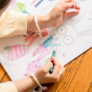 Può includere: Una persona sta colorando un coniglio dei cartoni animati con un pastello blu. Il coniglio indossa un abito e una cravatta. Il libro da colorare è aperto su un tavolo di legno. La persona indossa un braccialetto con perline bianche.