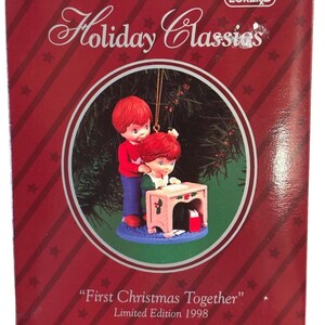 Puede incluir: Un adorno navideño con dos niños pelirrojos. Uno está de pie detrás del otro, que está sentado en un pequeño escritorio rosa. El adorno está sobre un fondo rojo con el texto "Holiday Classics" y "First Christmas Together" Edición Limitada 1998.