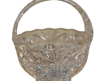 May include: A clear, cut-glass basket with a handle. The basket has a scalloped rim and is decorated with a repeating pattern of diamonds, circles, and floral designs. The basket sits on a small, round base.