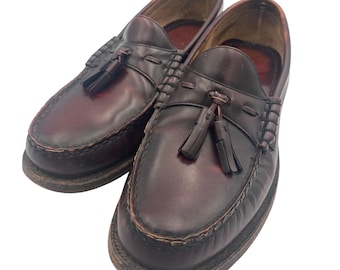 Pode incluir: Um par de mocassins de couro bordô com borlas decorativas. Os sapatos têm um design clássico com costuras visíveis ao redor das solas. O couro tem uma aparência ligeiramente desgastada.