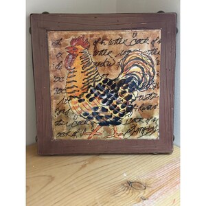 May include: A square decorative tile with a rooster illustration in orange, black, and brown. The tile is framed in a dark brown wooden border. Handwritten script surrounds the rooster.