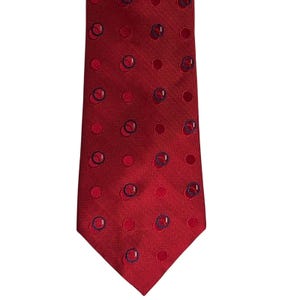May include: A red necktie with a repeating pattern of dark blue circles and red dots. The tie has a textured surface and a classic pointed end. This formal accessory is suitable for business or special occasions.