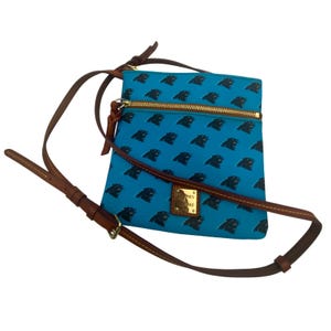 Puede incluir: Bolso bandolera azul con un patrón repetido del logotipo de pantera negra. El bolso tiene una cremallera dorada, una correa de cuero marrón y una pequeña etiqueta rectangular de la marca. El bolso mide aproximadamente 23 cm de ancho y 25 cm de alto.