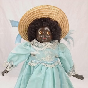 May include: A vintage porcelain doll with black hair and brown eyes, wearing a light blue dress with lace trim and a straw hat with a blue ribbon.