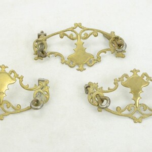 May include: Three ornate gold-toned metal drawer pulls with intricate scrollwork and a central oval design. The pulls have a vintage aesthetic and would be a great addition to a dresser, cabinet, or other furniture piece.