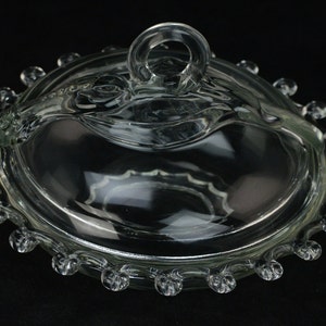 Puede incluir: Un plato de mantequilla de vidrio transparente con tapa y asa de pomo. El plato tiene un borde festoneado con pequeños pomos alrededor del borde.