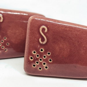 May include: A set of two red ceramic salt and pepper shakers with holes for dispensing. The shakers are shaped like a rectangle with rounded corners. The salt shaker has the letter "S" on it and the pepper shaker has the letter "P" on it.
