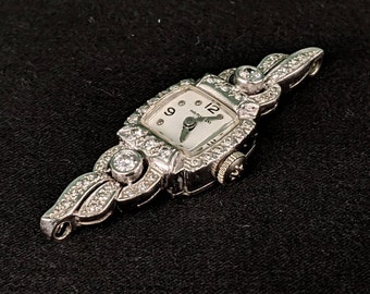 May include: A vintage wristwatch with a white face and diamond-encrusted band. The watch face has black Roman numeral hour markers and a black hand. The watch is set in a silver-toned metal.