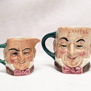 May include: Two ceramic creamers with green handles and painted faces. The larger creamer has the word "MICAWBER" written on the side.