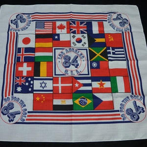 May include: A white bandana with a blue border and red stripes. It features a grid of flags from different countries, including the United States, Canada, Japan, and the United Kingdom. The center of the bandana has a circle with the text "Los Angeles 84 U.S.A." and the corners have the text "Go For The Gold 84".