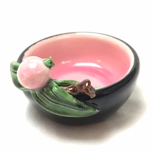 May include: A small black ceramic bowl with a pink interior and a 3D floral design. The bowl has a pink flower with green leaves and a brown stem.