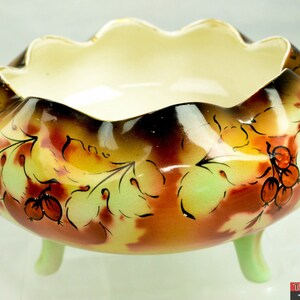 Puede incluir: Un cuenco con pie con un borde festoneado y un diseño floral. El cuenco está decorado con hojas verdes, marrones y naranjas y bayas rojas. El cuenco está hecho de cerámica y tiene un acabado brillante.