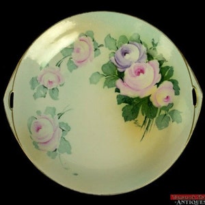 May include: A vintage porcelain serving dish with two handles. The dish is decorated with a hand-painted floral design featuring pink, purple, and white roses with green leaves.