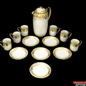 Op de afbeelding: Een vintage theeservies met een witte theepot en zes kopjes en schoteltjes. De set heeft een gouden en zwart bloemenmotief.