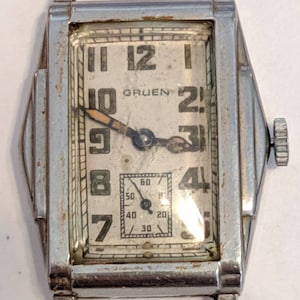 May include: A silver-toned Gruen wristwatch with a rectangular face and black Roman numeral hour markers. The watch has a small second hand dial at the bottom of the face.