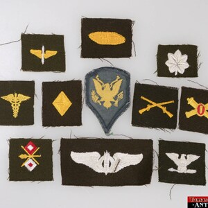 May include: A collection of 11 military patches on a green background. The patches are embroidered with various symbols, including a medical caduceus, a compass, a eagle, crossed rifles, artillery, and an airplane.