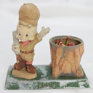 May include: A vintage Elmer Fudd match holder featuring a cartoon Elmer Fudd figurine holding a rifle and a matchstick holder shaped like a tree stump. The base is metal and has the word "ELMER" printed on it.