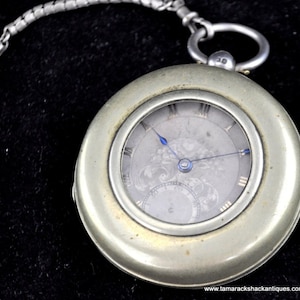 May include: Silver pocket watch with a white face and blue hands. The watch has a decorative design on the face and a chain attached to the top.