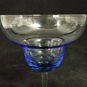Op de afbeelding: Een helder glazen margarita-glas met een blauwe rand en voet. Het glas is leeg en staat op een zwart oppervlak.