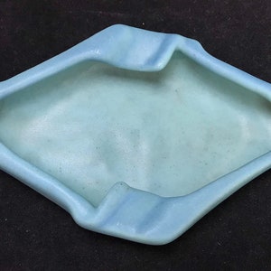 Op de afbeelding: Een lichtblauwe asbak van keramiek met een uniek, asymmetrisch ontwerp. De asbak heeft een brede, ondiepe kom en een verhoogde rand.