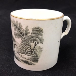 May include: White ceramic mug with a black and white landscape scene printed on the side. The mug has a gold rim.