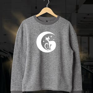 Könnte beinhalten: Graues Sweatshirt mit weißem Mond- und Katzenmotiv. Das langärmlige Oberteil hat einen Rundhalsausschnitt und gerippte Bündchen und Saum. Die Katze sitzt im Mond mit Sternen. Das Sweatshirt hängt an einem Holzbügel.