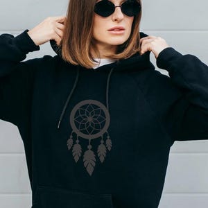 Può includere: Felpa con cappuccio nera con un design acchiappasogni grigio scuro. L'acchiappasogni ha una rete circolare con un motivo floreale e tre piume pendenti. La felpa ha un cordino e un cappuccio.