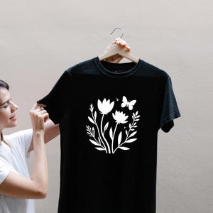 Puede incluir: Camiseta negra con un diseño floral blanco que presenta dos flores, hojas y una mariposa. El gráfico está centrado. La camiseta está colgada en una percha de madera.