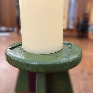 May include: A cylindrical off-white pillar candle sits atop a small, round, green-painted wooden stool with three legs. The stool features a visible wood grain texture under the green paint, and the candle has a smooth, matte surface finish.