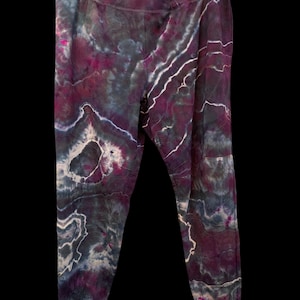 Pode incluir: Um par de leggings tie-dye em tons de roxo, preto e branco. As leggings têm um padrão marmorizado com linhas brancas semelhantes a veias. O tecido parece macio e elástico. As leggings estão penduradas num cabide preto.
