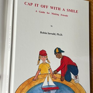 May include: A hardcover book titled "CAP IT OFF WITH A SMILE: A Guide for Making Friends" by Robin Inwald, Ph.D. The cover features a colorful illustration of two children and a sailboat on a rainbow.