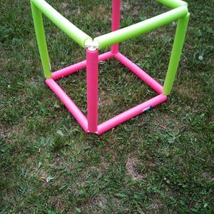 Puede incluir: Estructura cúbica hecha de fideos de piscina de espuma rosa y verde, colocada sobre hierba verde. Los cuadrados superior e inferior son rosas, con soportes verticales verdes.