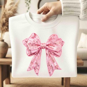 May include: White sweatshirt featuring a large pink sequined bow. The bow has a detailed design with a darker pink outline and is centered on the front of the shirt. The background has a subtle chevron pattern.