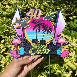 Op de afbeelding: Een kleurrijke taarttopper met een tropisch thema. Het heeft een gouden "40" en de naam "Ellie" in script. Roze flamingo's, palmbomen en vliegtuigen staan tegen een zonsondergang achtergrond. De topper wordt door een hand vastgehouden.