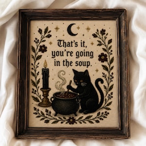 Könnte beinhalten: Ein gerahmtes Stickbild auf beigem Stoff zeigt eine schwarze Katze, die in einem dampfenden Kessel rührt. Der Text lautet, That's it, you're going in the soup. Eine schwarze Kerze, eine Mondsichel, Sterne und florale Ranken umrahmen das Motiv.