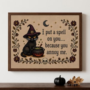 Könnte beinhalten: Ein gerahmtes Kreuzstich-Stickbild auf beigem Stoff mit einer schwarzen Katze in einem violetten Hexenhut neben einem Kessel. Der Text lautet, I put a spell on you... because you annoy me. Das Design umfasst eine Mondsichel, Sterne, einen Totenkopf und florale Bordüren.