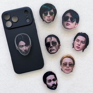 May include: A matte black smartphone case with a circular portrait sticker of a person wearing sunglasses attached to the back. Six additional circular portrait stickers of various individuals wearing sunglasses are arranged around the phone case.