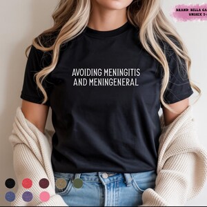 Op de afbeelding: Zwart unisex T-shirt met de witte tekst "AVOIDING MENINGITIS AND MENINGENERAL". Het T-shirt is van Bella Canvas 3001. Het model draagt een blauwe jeans en een crème vest.