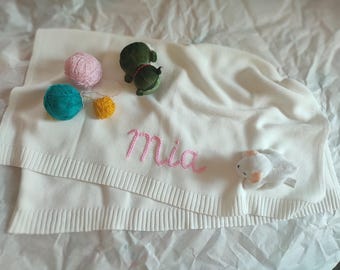 May include: A white knitted baby blanket with the name "Mia" embroidered in pink. The blanket is decorated with small plush toys and colorful yarn balls. A soft and cozy item for a baby.