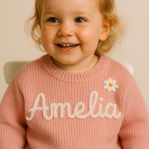 May include: A pink knitted sweater with the name "Amelia" in white script across the front. A small white daisy is embroidered near the right shoulder. The sweater is worn by a smiling child.