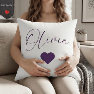 May include: A square white throw pillow featuring the name Olivia written in a dark purple cursive script font. Below the name is a solid dark purple heart shape. The pillow has a clean, simple design with no other patterns or textures visible.