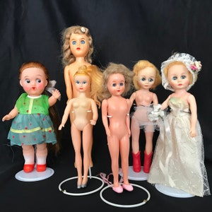 Op de afbeelding: Zes vintage plastic poppen met blond, bruin en zwart haar. De poppen dragen verschillende outfits, waaronder een groene jurk, een witte jurk, een blauwe jurk en een roze jurk. De poppen staan op een witte achtergrond.