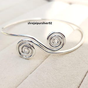 Op de afbeelding: Een zilverkleurige manchetarmband met een spiraalvormig ontwerp aan beide uiteinden. De armband heeft een gladde, gepolijste afwerking en de tekst "shrejaipursilver92". De spiralen geven de armband een unieke uitstraling.