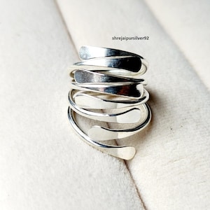 May include: A silver-colored wrap ring with a spiral design. The ring features a series of curved, leaf-shaped elements that overlap and coil around the finger. The ring is made of a shiny, reflective metal. The text "shrejaipursilver92" is visible.