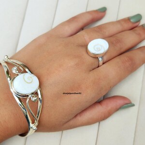 Op de afbeelding: Een zilveren armband- en ringenset. De armband heeft een gebogen ontwerp met een grote ovale witte steen met een spiraalpatroon. De ring heeft een bijpassende witte steen. De sieraden worden op een hand getoond.