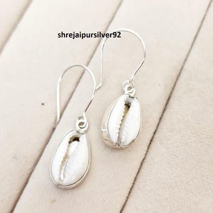 Puede incluir: Pendientes de plata con conchas de cauri blancas. Las conchas están engastadas en plata y cuelgan de ganchos de plata. El texto "shrejaipursilver92" es visible.